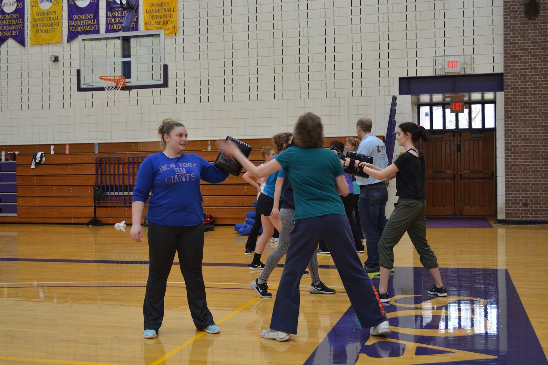 People practicing stage combat in a gymnasium. One woman holds a prop, while others are armed with training weapons.