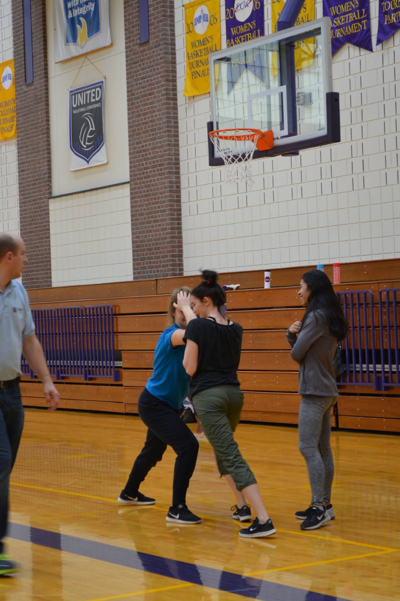 Two women practicing self-defense in a gymnasium, another woman watches. A man stands to the left, observing.