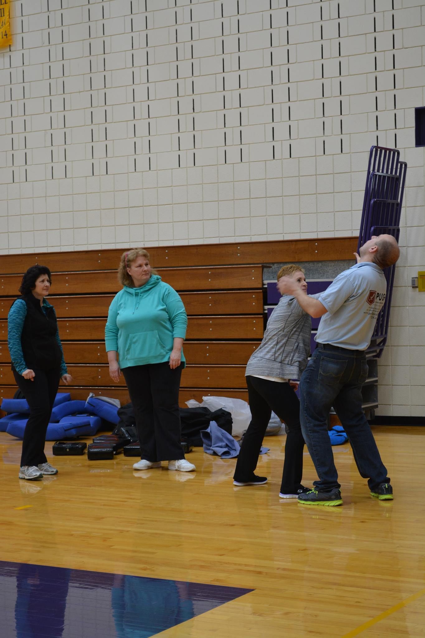 Four people in a gymnasium; two women watch as a man demonstrates a defensive move on another woman.