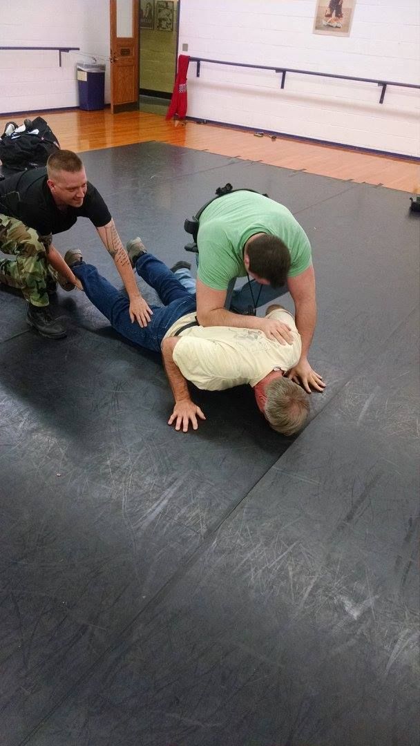 Three men practicing a grappling technique on a mat. One man is on the ground, face down, while another kneels on his back. The third man assists by holding the legs.