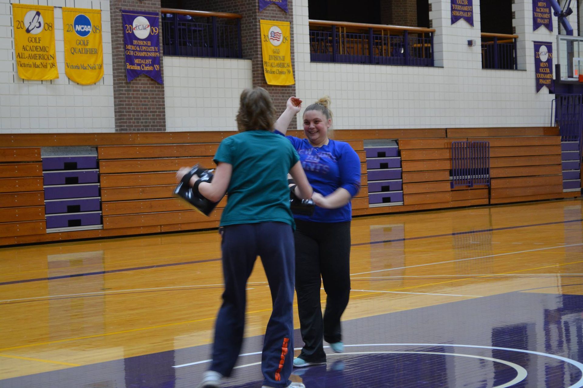 Two women high-fiving in a gymnasium. One wears a green shirt and black gloves, the other a blue shirt and is smiling.