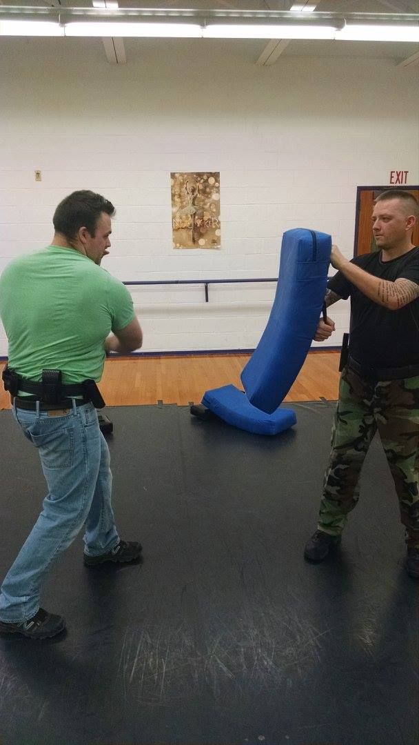 Two men practicing self-defense; one in a green shirt punches a blue padded training bag held by the other man in camouflage pants.