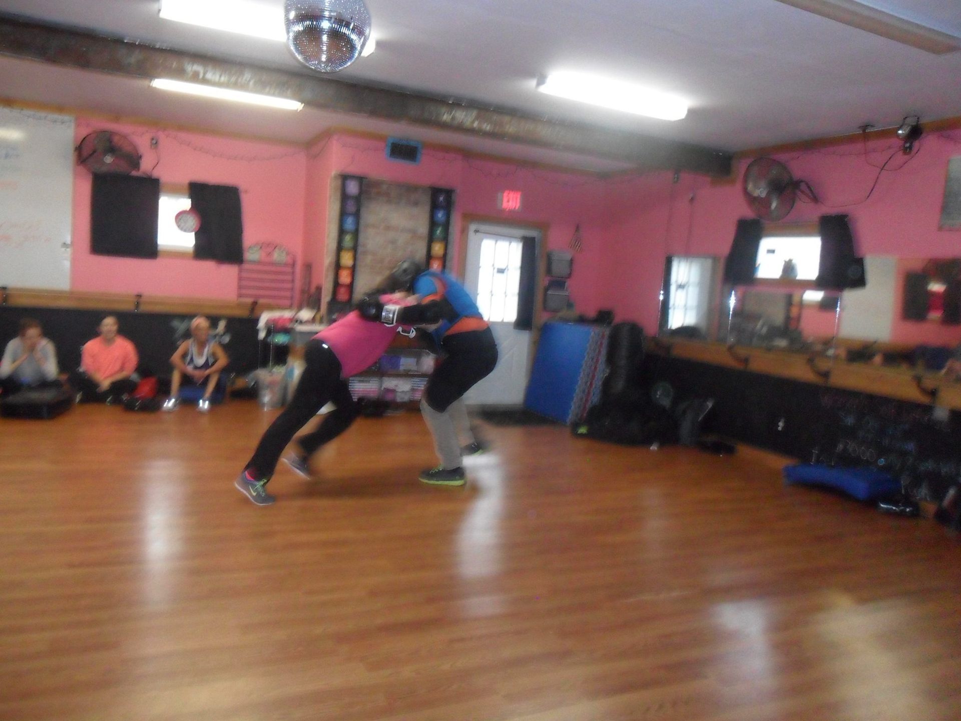 Two people wrestling in a pink gym. One in pink leans forward, the other in blue holds them. Others watch from the background.