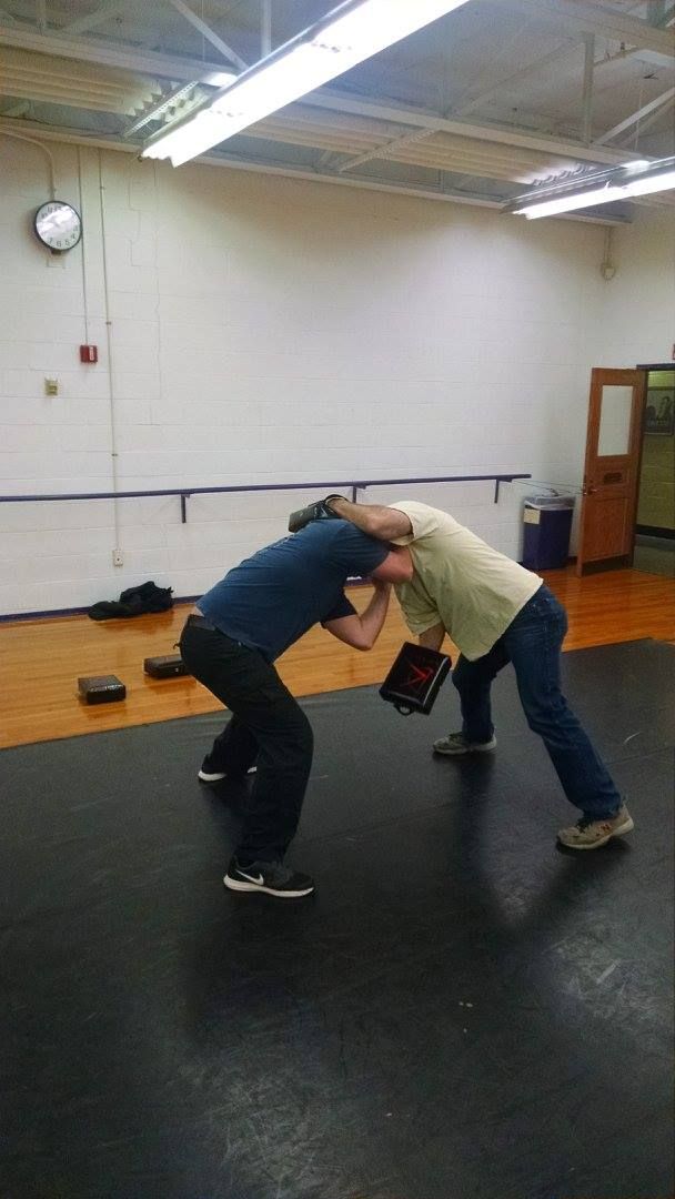 Two people wrestling on a mat in a gym. One in a blue shirt and the other in a tan shirt are locked in a headlock.