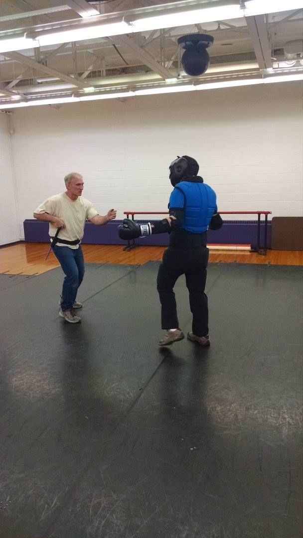 Two people practicing martial arts in a gym. One person wears protective gear, the other is lightly dressed.