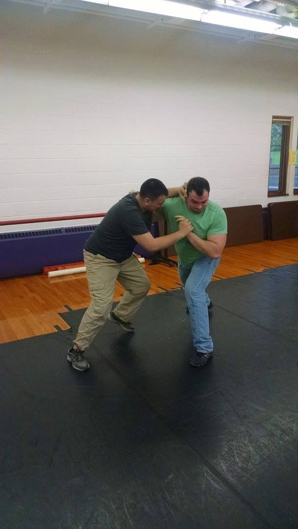 Two men in a gym practice a self-defense move. One in black shirt and khaki pants prepares to throw the other, in a green shirt and jeans.