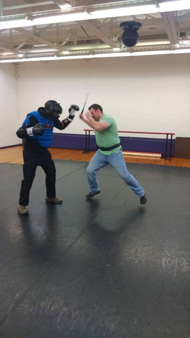 Two men in a gym practicing sword fighting. One in protective gear; the other in casual clothing, wielding a sword.