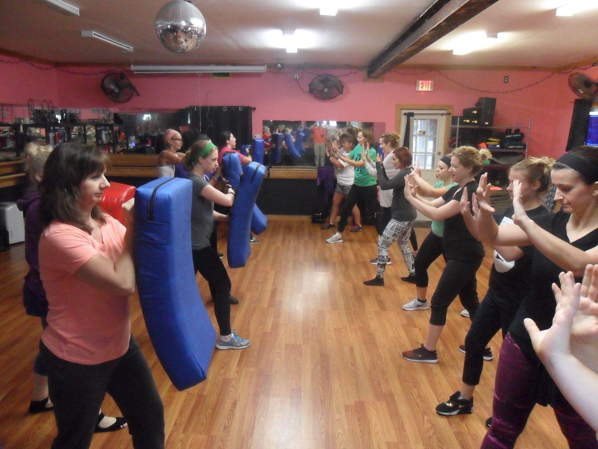 A group of women practice self-defense techniques in a pink-walled studio. Some hold blue pads, others practice punches.