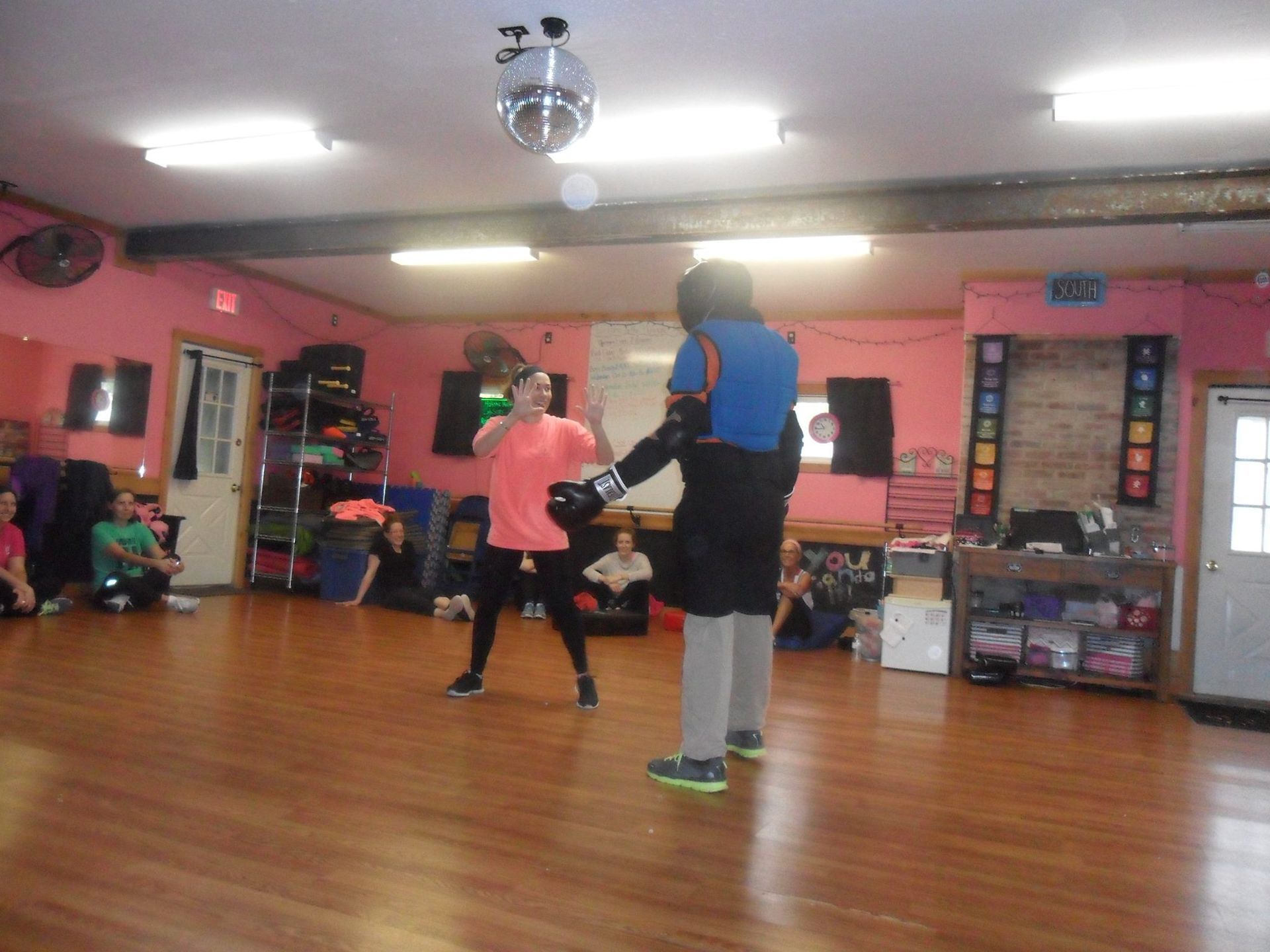 Two people sparring in a dance studio, one wearing protective gear, the other throwing a punch. Pink walls, wooden floor, and spectators.