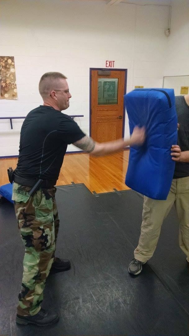 A man in camouflage pants punches a blue pad held by another person indoors. The man is wearing a black shirt.