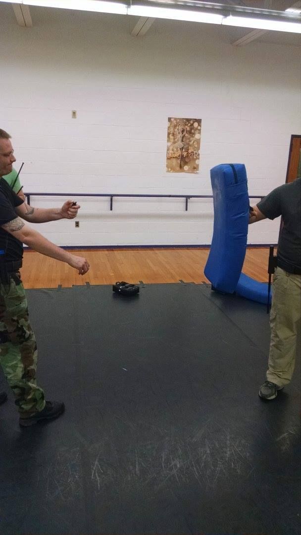 A man in camouflage pants practices self-defense, throwing a punch at a training pad held by another man in a gym.