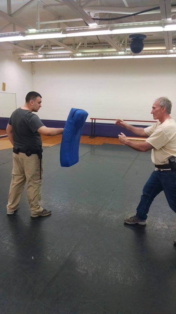 Two men practicing defensive tactics in a gymnasium. One holds a blue striking shield; the other practices a punch or strike.