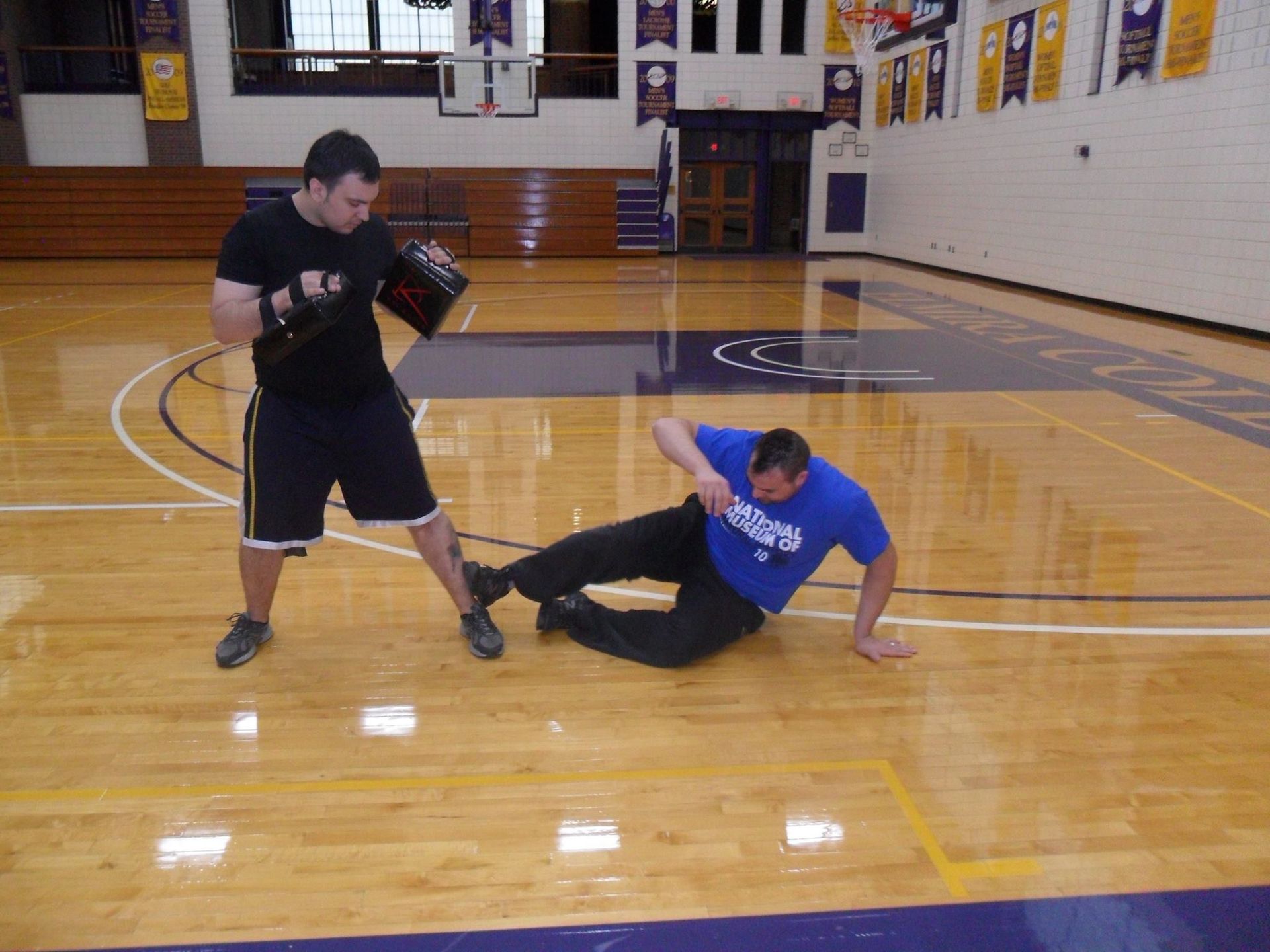 Two men in a gym: one in black shorts and shirt holding a pad, the other in blue, on the floor, performing a self-defense exercise.