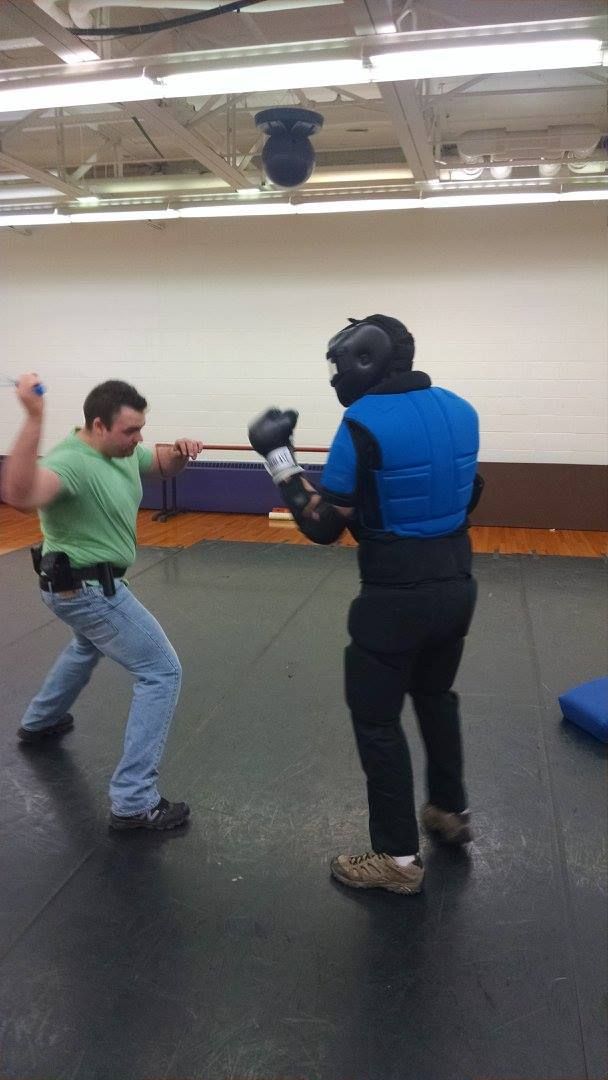 Man in green shirt and jeans swings a knife at person in protective gear inside a gym.