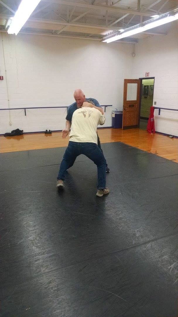 Two men in a wrestling stance on a mat. One wears a tan shirt and jeans, the other a blue shirt. The setting is a gym with a wooden floor.