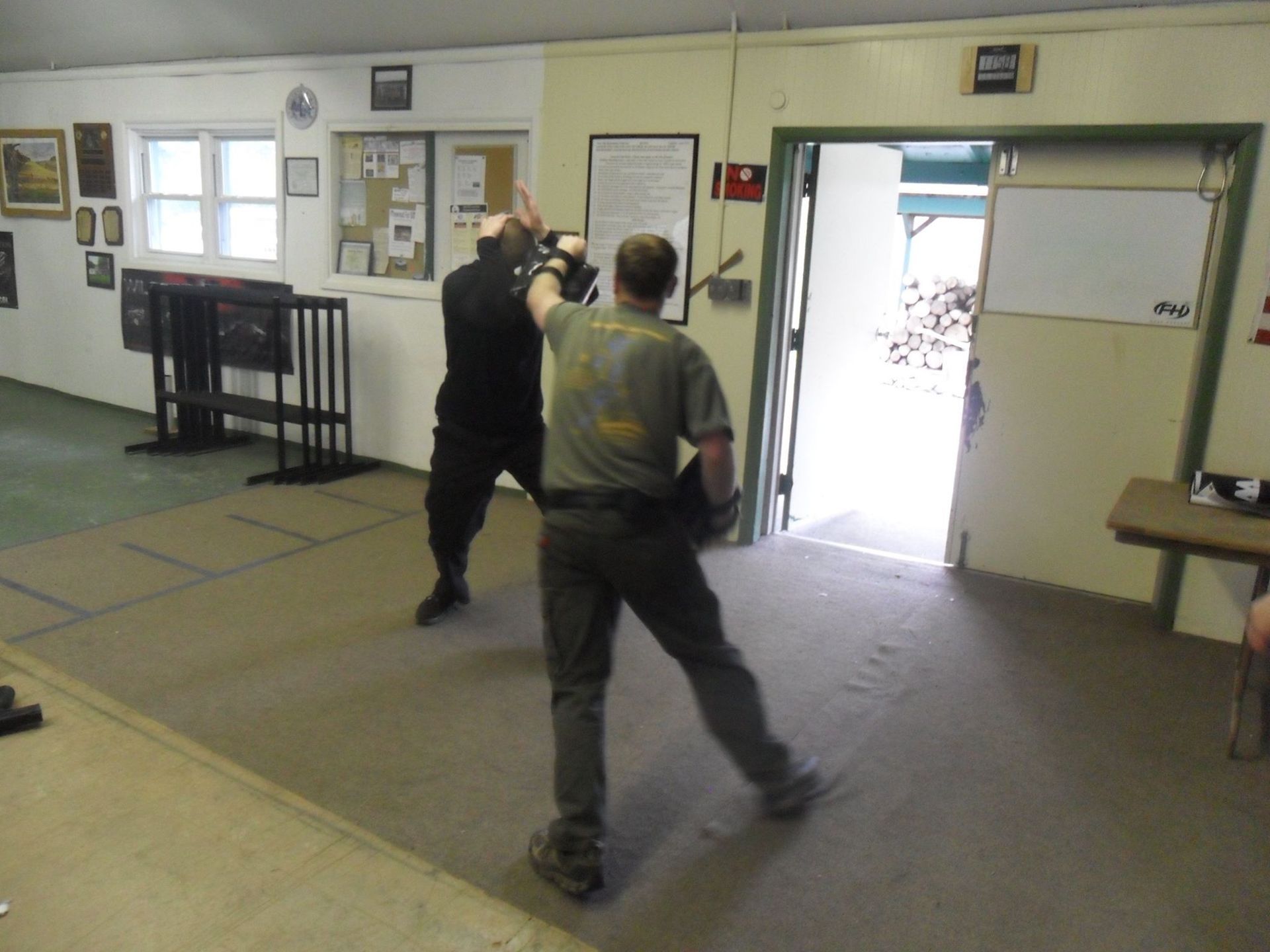 Two men practice martial arts in a training room. One, in dark clothing, blocks an overhead strike from the other, in a green shirt.