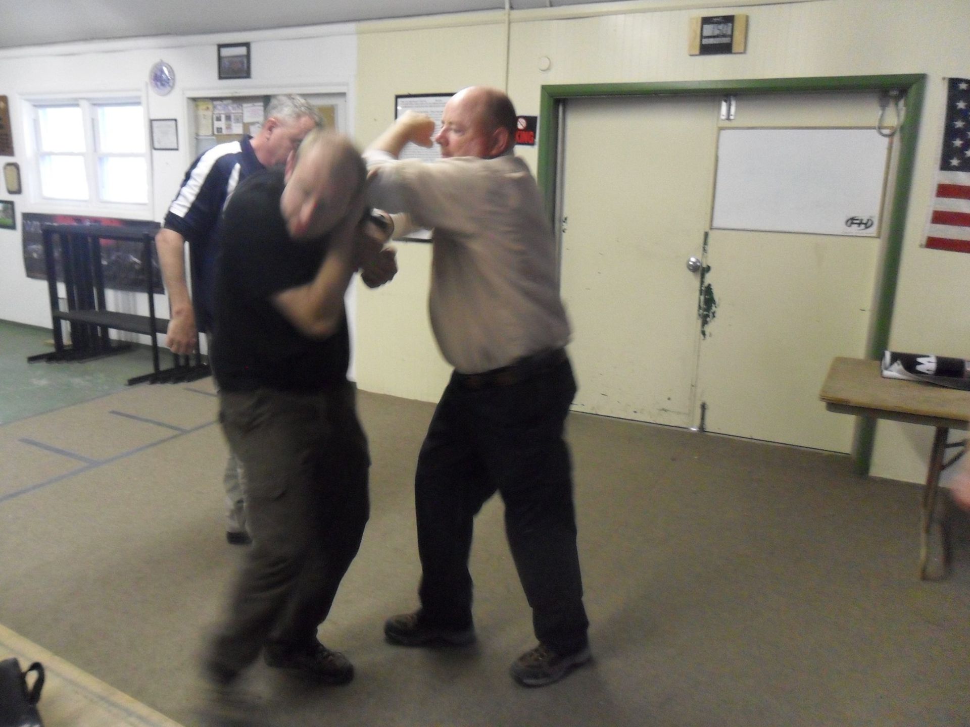 Two men in a room practicing self-defense; one throws a punch at the other's head. A third man watches nearby.