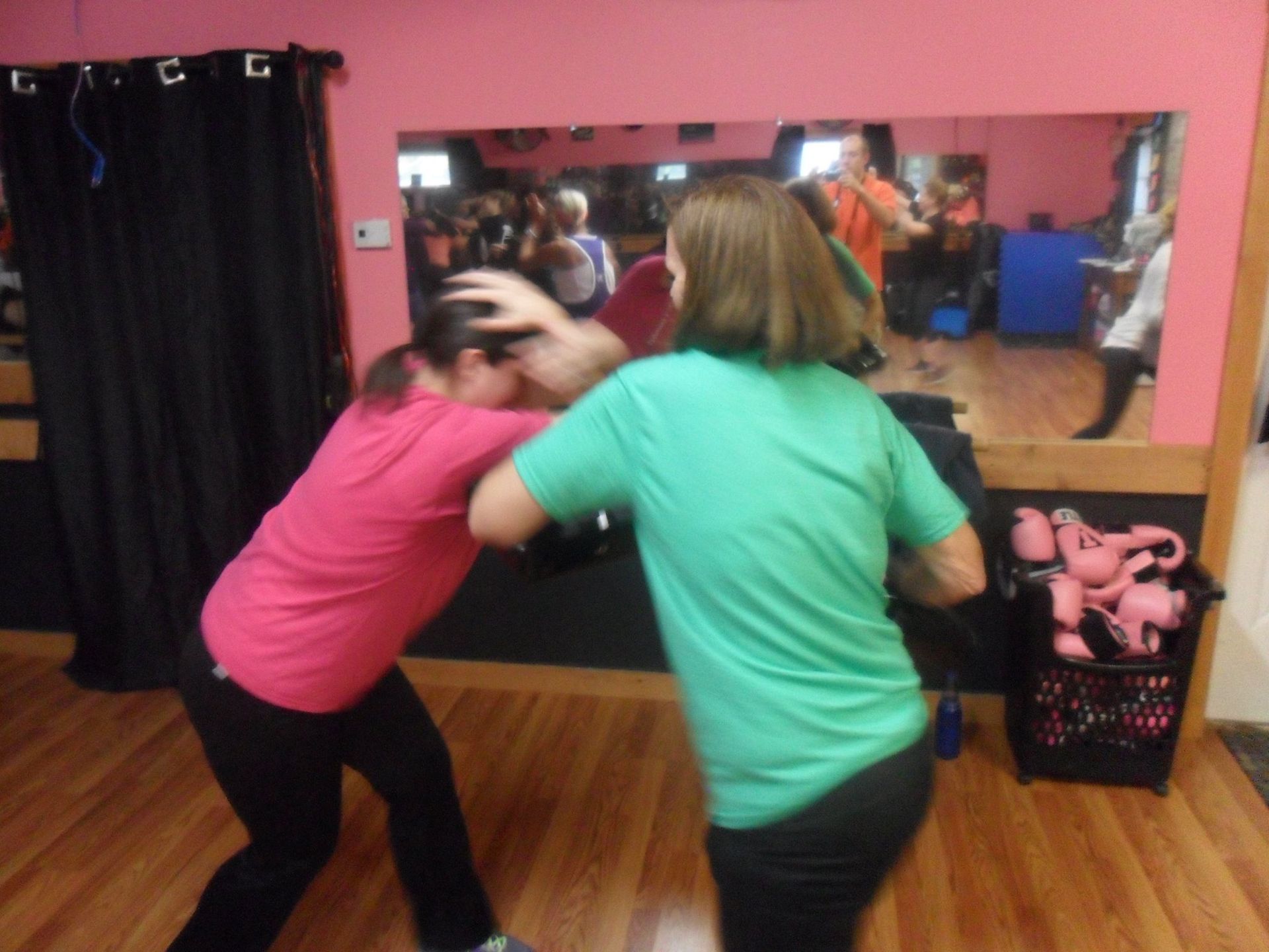 Two women in a gym practice self-defense, with one striking the other who is blocking her head.  A mirror reflects other people and equipment.