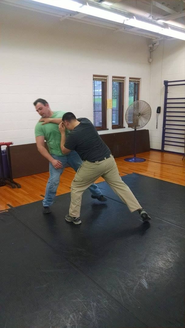 Two men practicing self-defense in a gym. One man in green grabs the other man in black. The other man blocks with an arm and steps to the side.