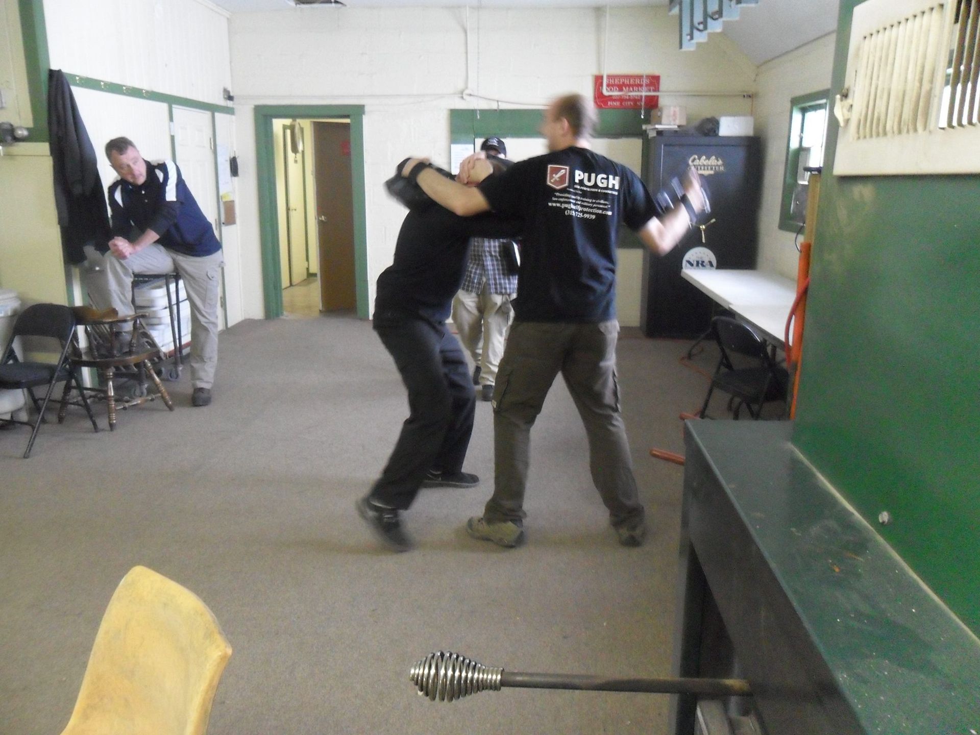 Two men in a room practicing self-defense. One is grabbing the other's shoulders; another man watches in the background.