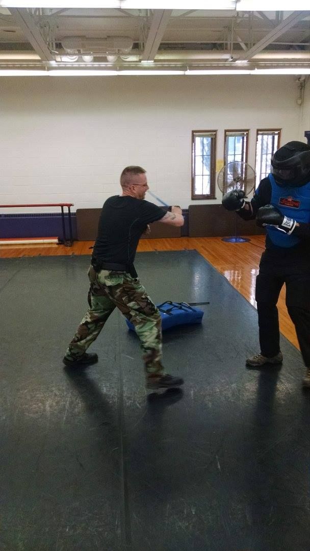 Man in camouflage pants and black shirt practices knife fighting with a person in protective gear indoors.