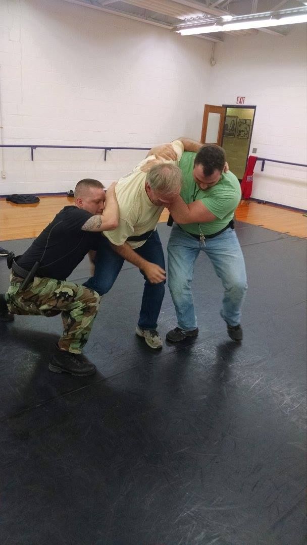 Three men practicing a grappling technique on a mat. One man is in the center, while two others hold him by his arms.