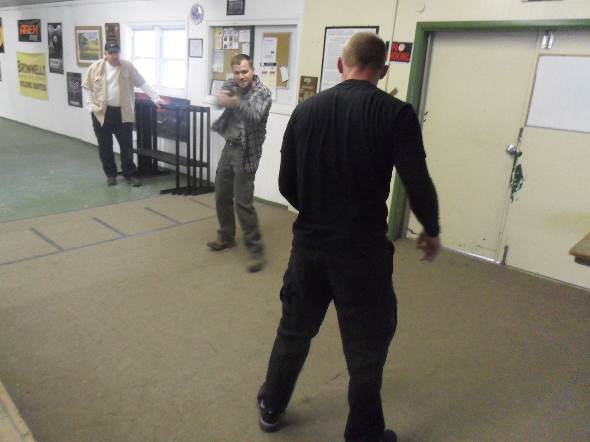 Three men in a room. One aims a gun, another stands behind him, and a third stands farther away.
