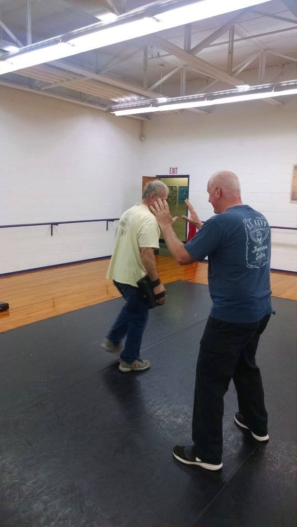 Two men practice self-defense in a gym. One blocks while the other moves away. Wooden floor, blue and yellow shirts.