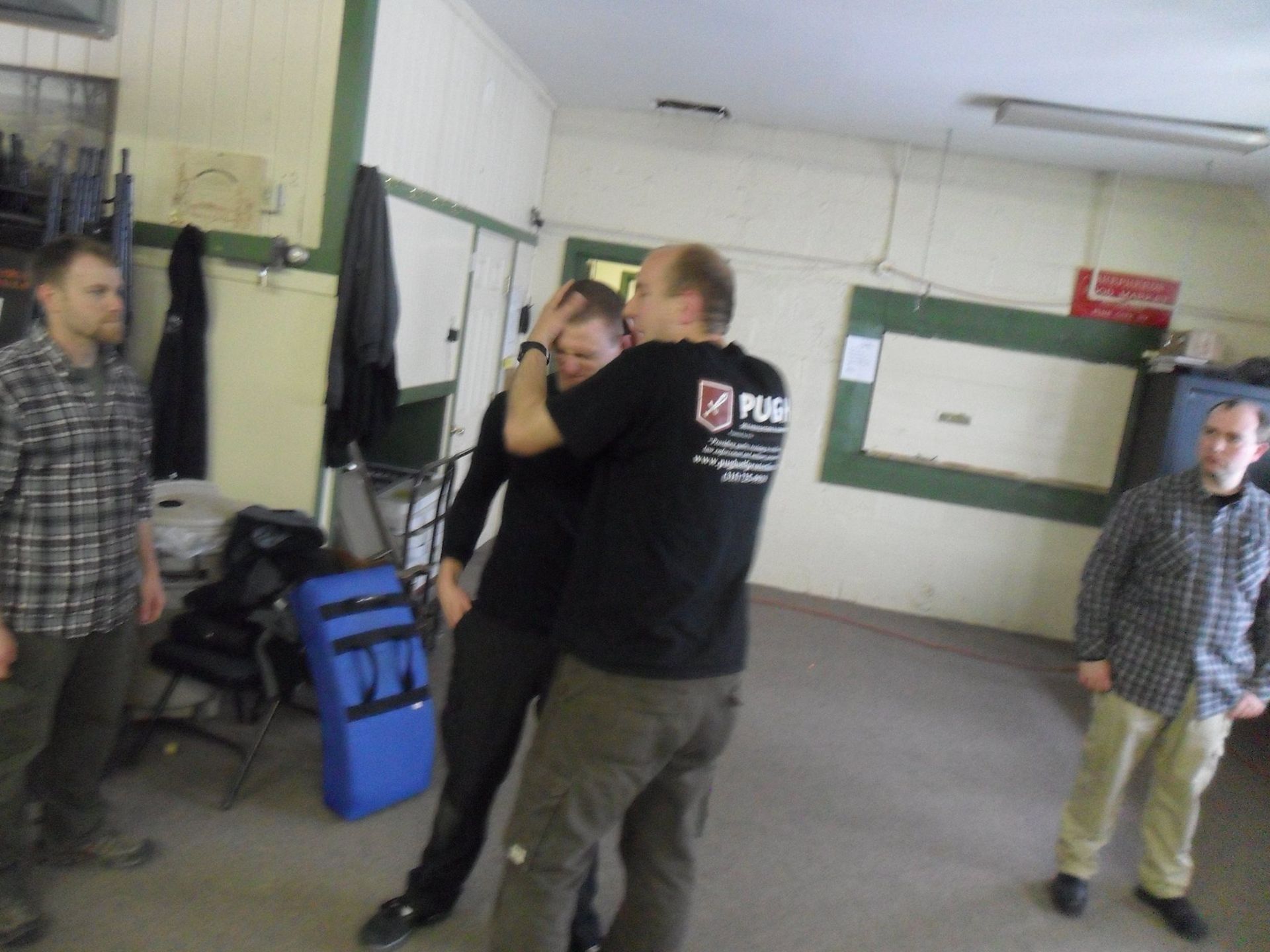 Two men in a room practicing self-defense. One grips the other's head, with others watching. Indoors with neutral walls.