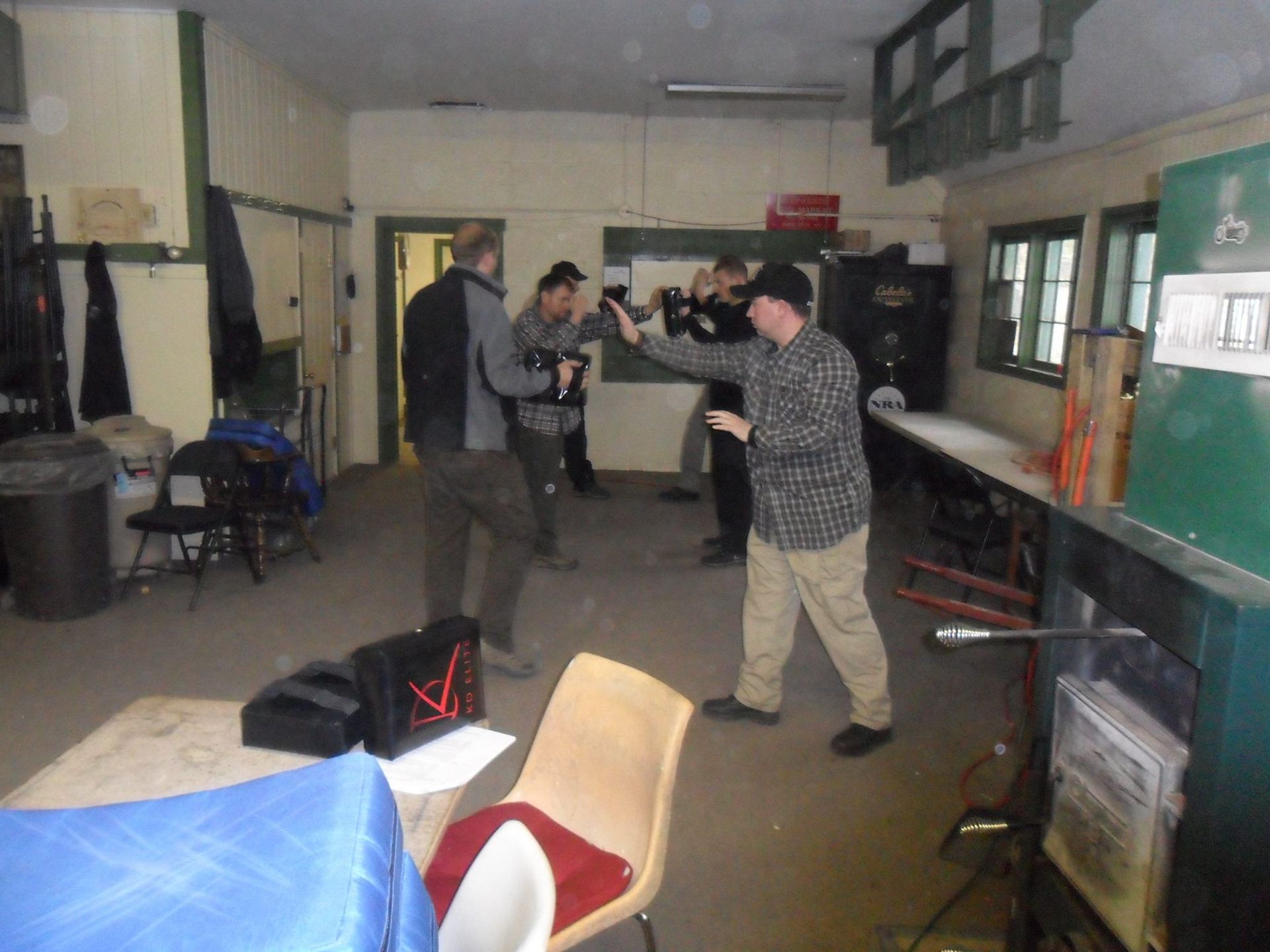 Men practicing martial arts in a room. Some are holding mock weapons. The setting appears to be a workshop or training space.