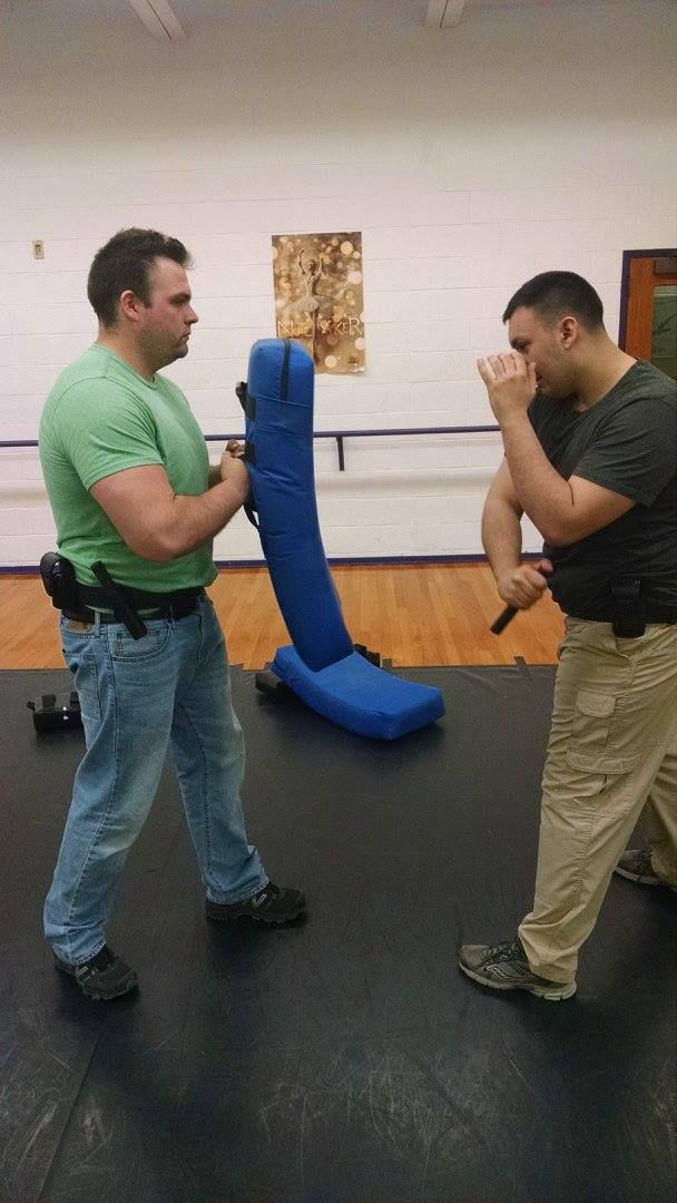 Two men in an indoor space training. One holds a blue padded shield while the other practices a defensive stance, holding a baton.