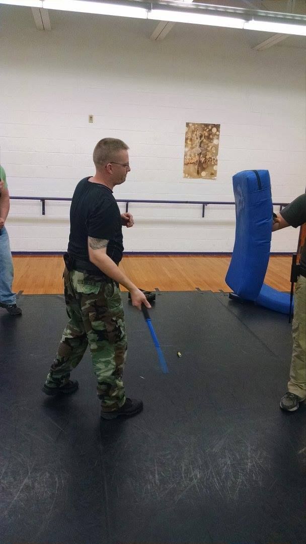 Man in camo pants and black shirt practices baton strikes on a blue padded training dummy indoors.
