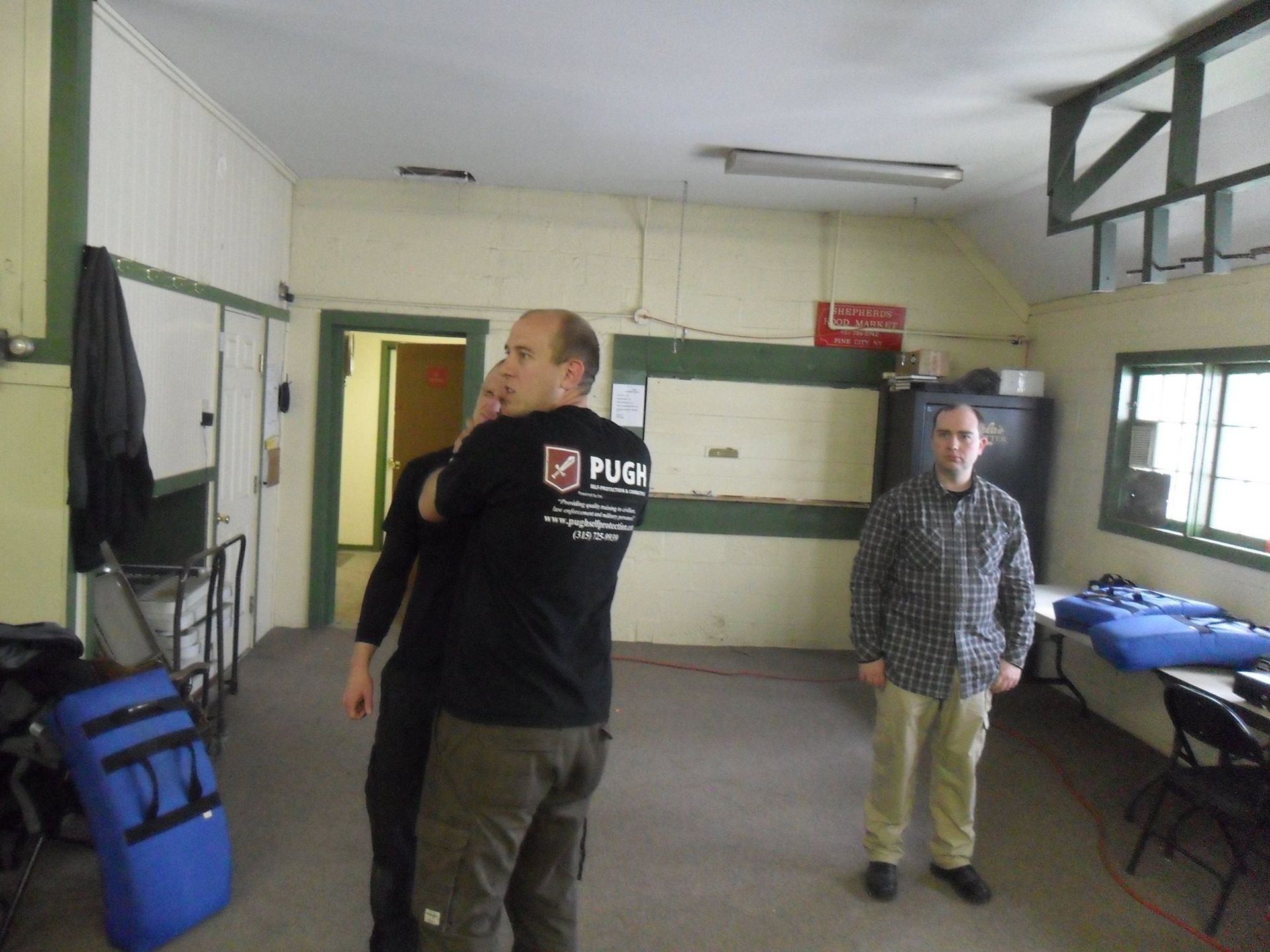 Man in black shirt demonstrates a neck hold on another man, while a third man watches in a sparsely furnished room.