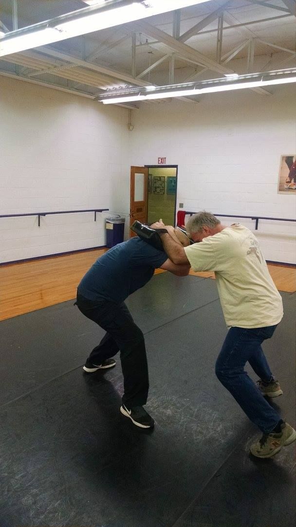 Two men wrestling on a black mat in a gymnasium. One man is bent over while the other has his arm around his neck.