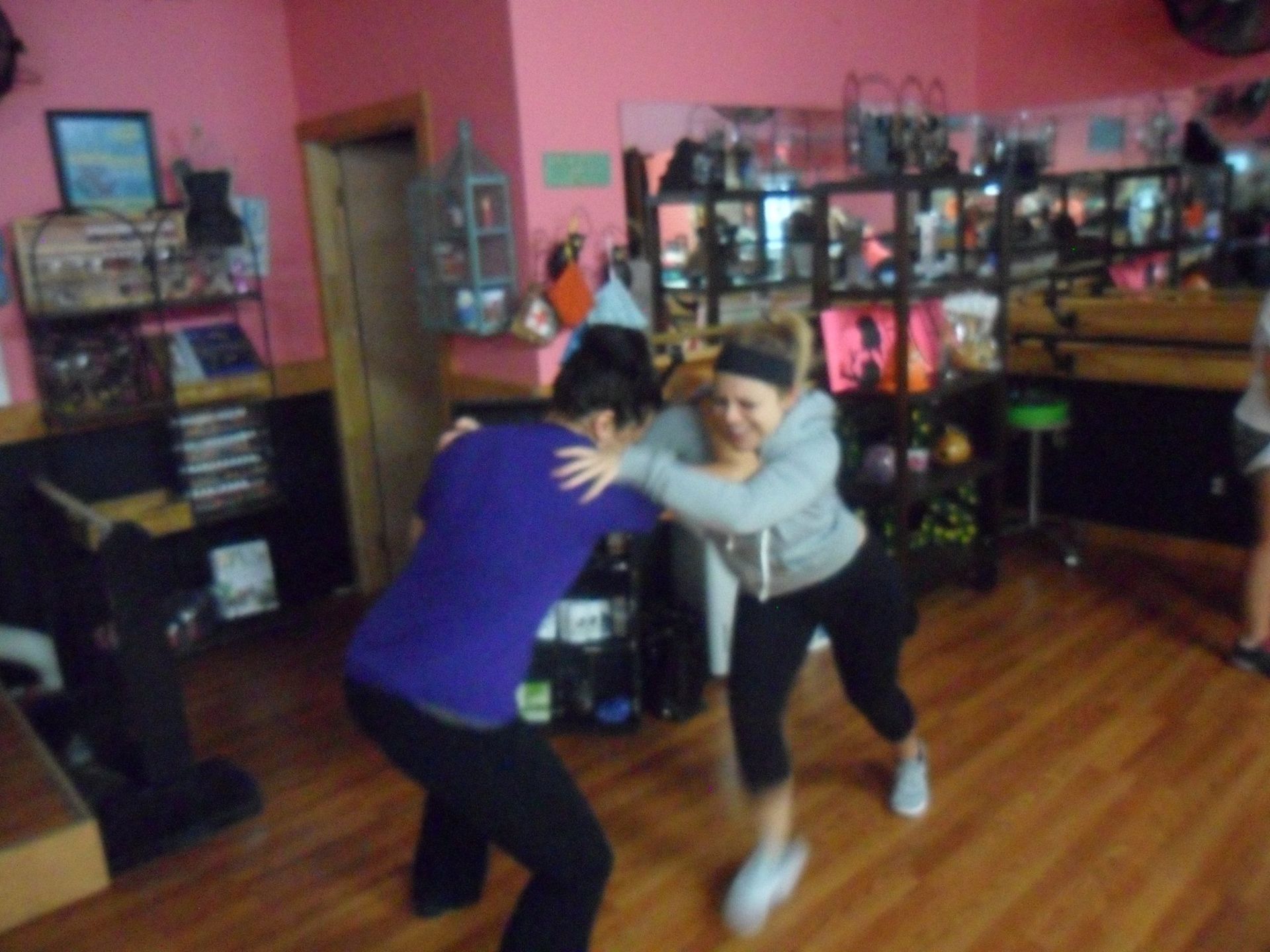 Two women in a pink-walled studio are playfully wrestling. One wears purple, the other gray, both smiling.