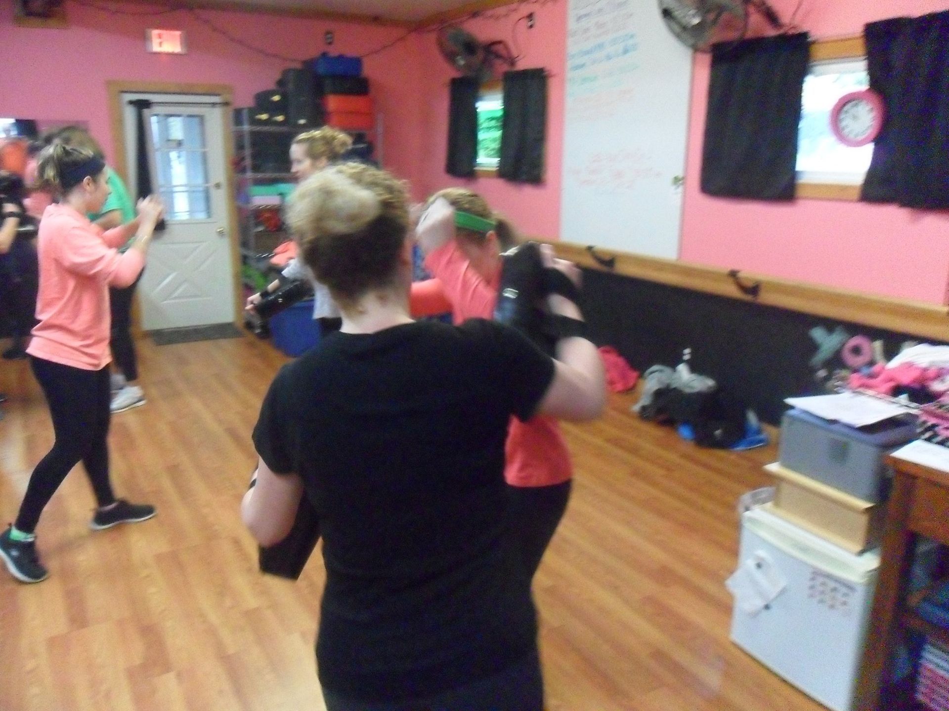 People exercising in a gym setting. Several are doing boxing moves, wearing workout clothes. Pink walls and wooden floors are visible.