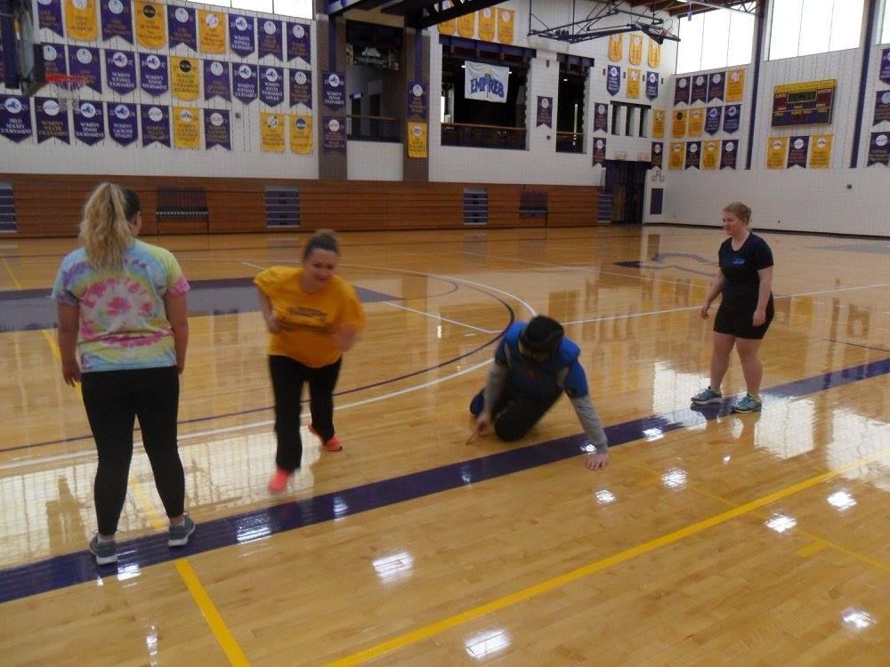 Four people in a gymnasium play a game on the floor. One kneels, reaching for something, while others watch. Banners hang on the walls.