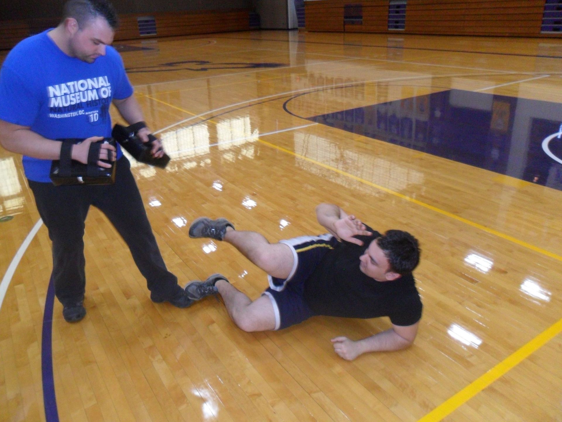 A man on a gym floor practicing self-defense, while another holds a pad. The man is on his side, blocking with one arm.