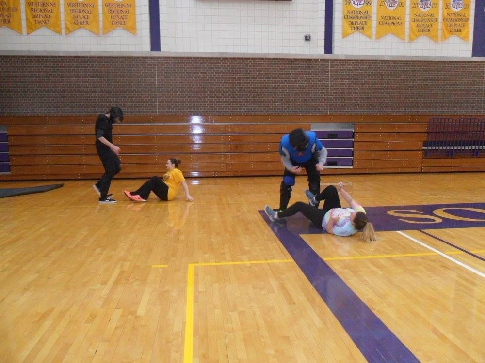 Four people in a gymnasium practicing martial arts. Two are on the floor, while the other two are standing, with one appearing to instruct.