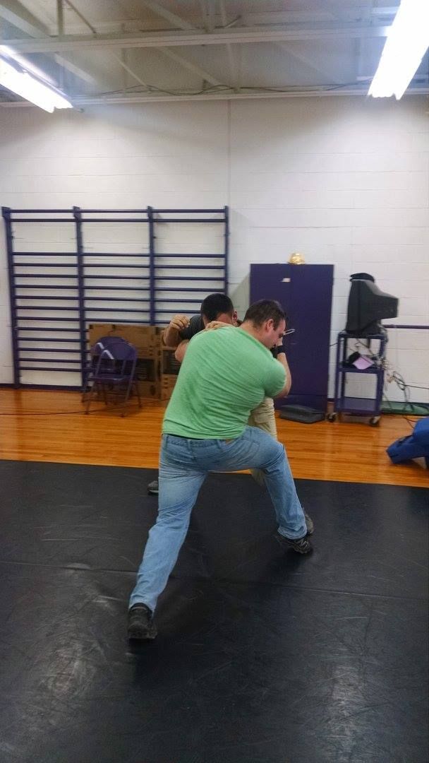 Man in green shirt and jeans wrestling with another person on a mat in a gym.