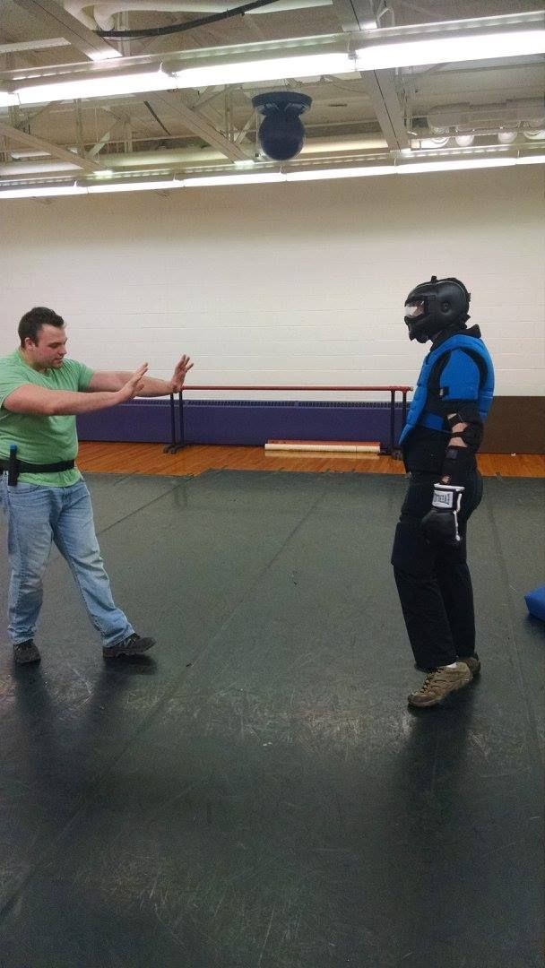 A man in a green shirt blocks a person in protective gear during a martial arts training session in a gym.