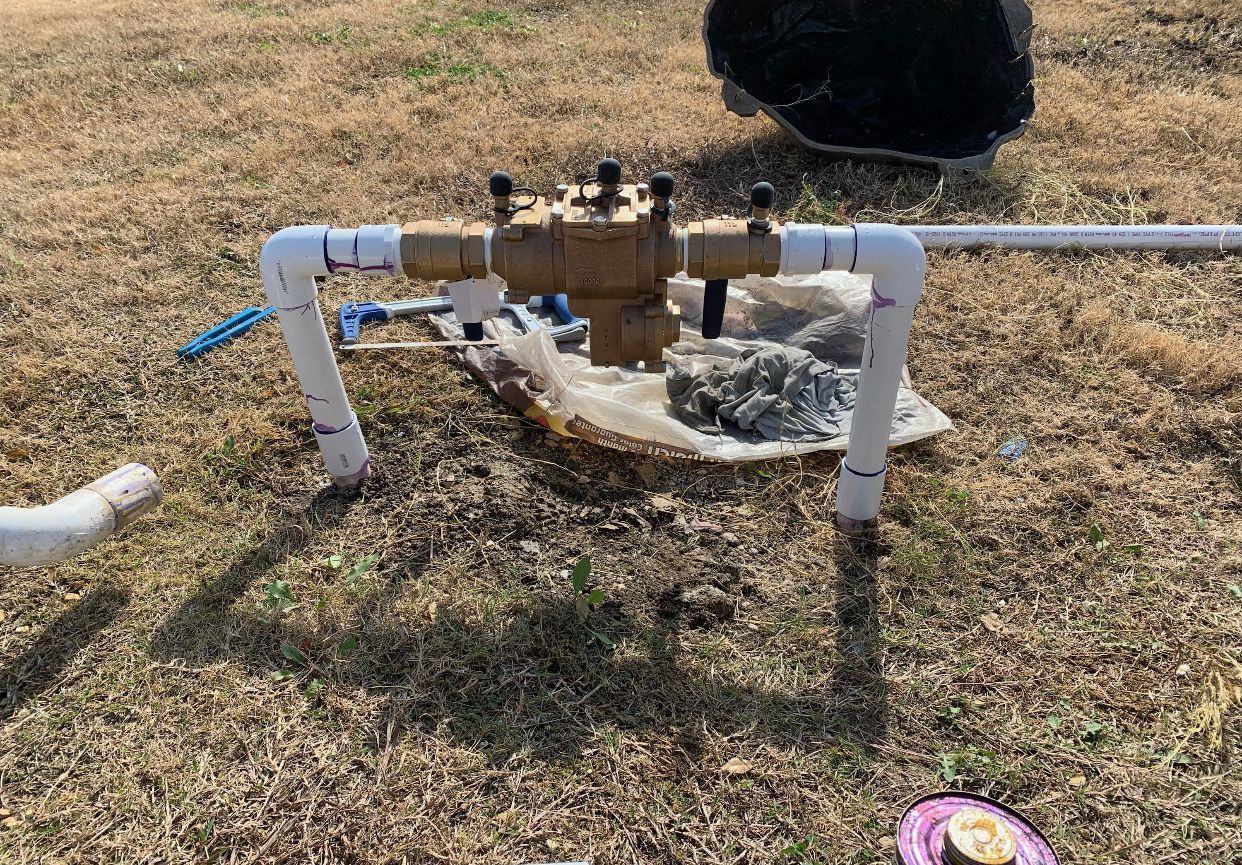 A sprinkler system backflow preventer with PVC pipes, sitting on dry grass.