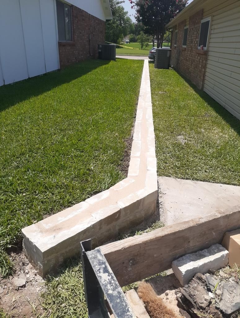 Concrete walkway between two buildings, partially constructed with unfinished edges. Green grass surrounds.