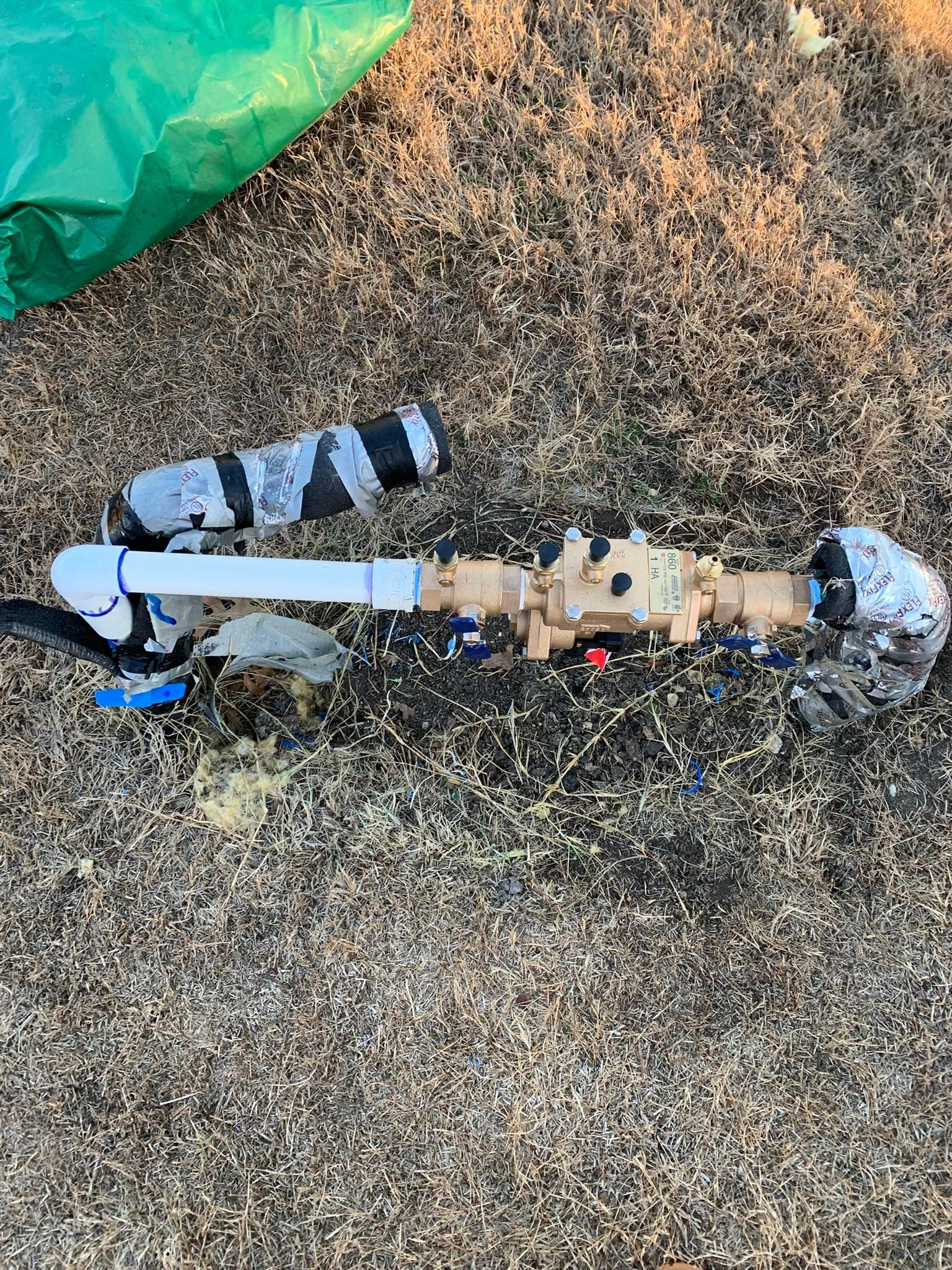 Irrigation system components on gravel ground. Gray and black pipes, brass valve. Green trash bag in background.