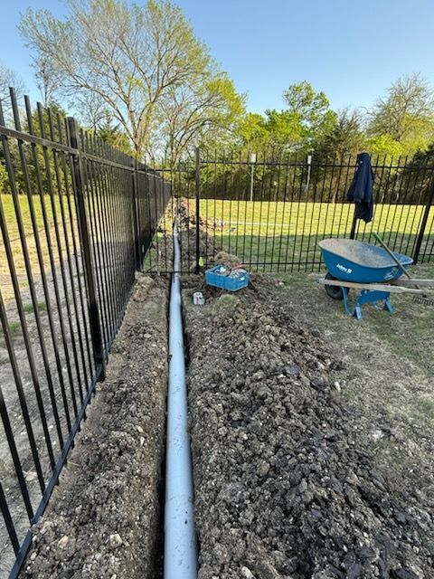 Trench with gray pipe next to black fence, blue wheelbarrow, blue box, and dirt, outdoors.