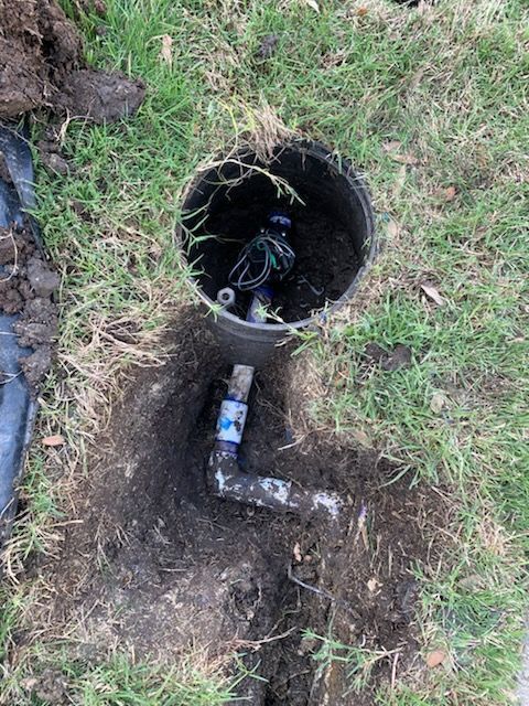 Open sprinkler valve box, pipe fittings, and buried wires in grass.