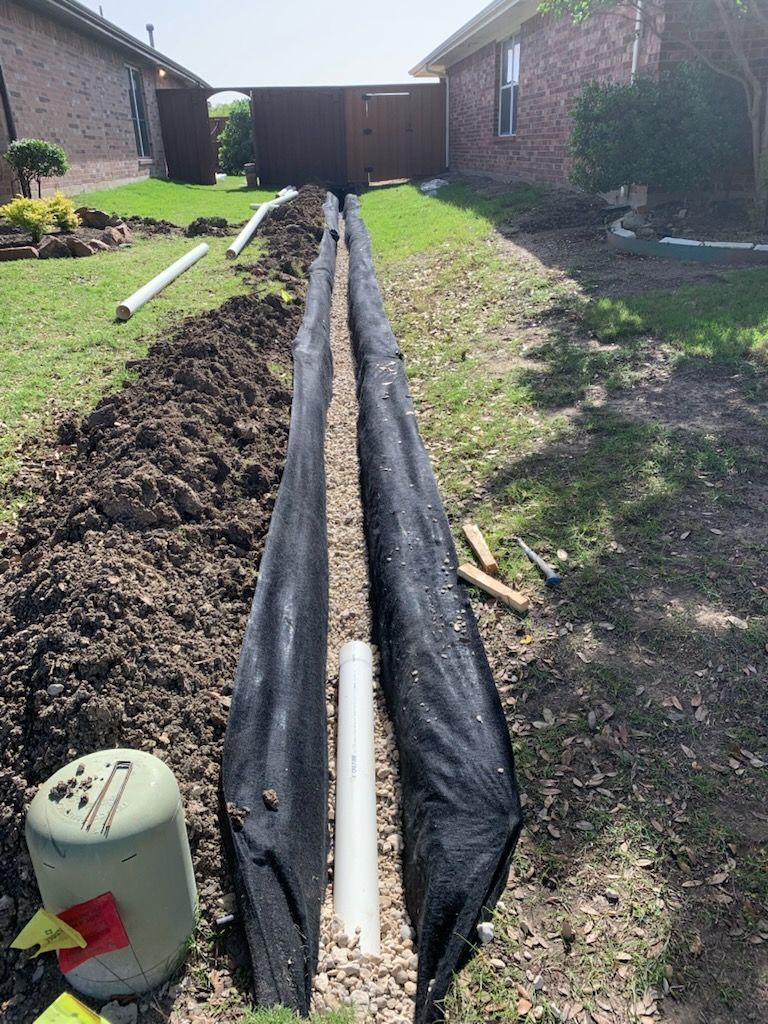 Construction of a drainage system in a yard. Black fabric lines the trench with gravel, and a white pipe is visible.