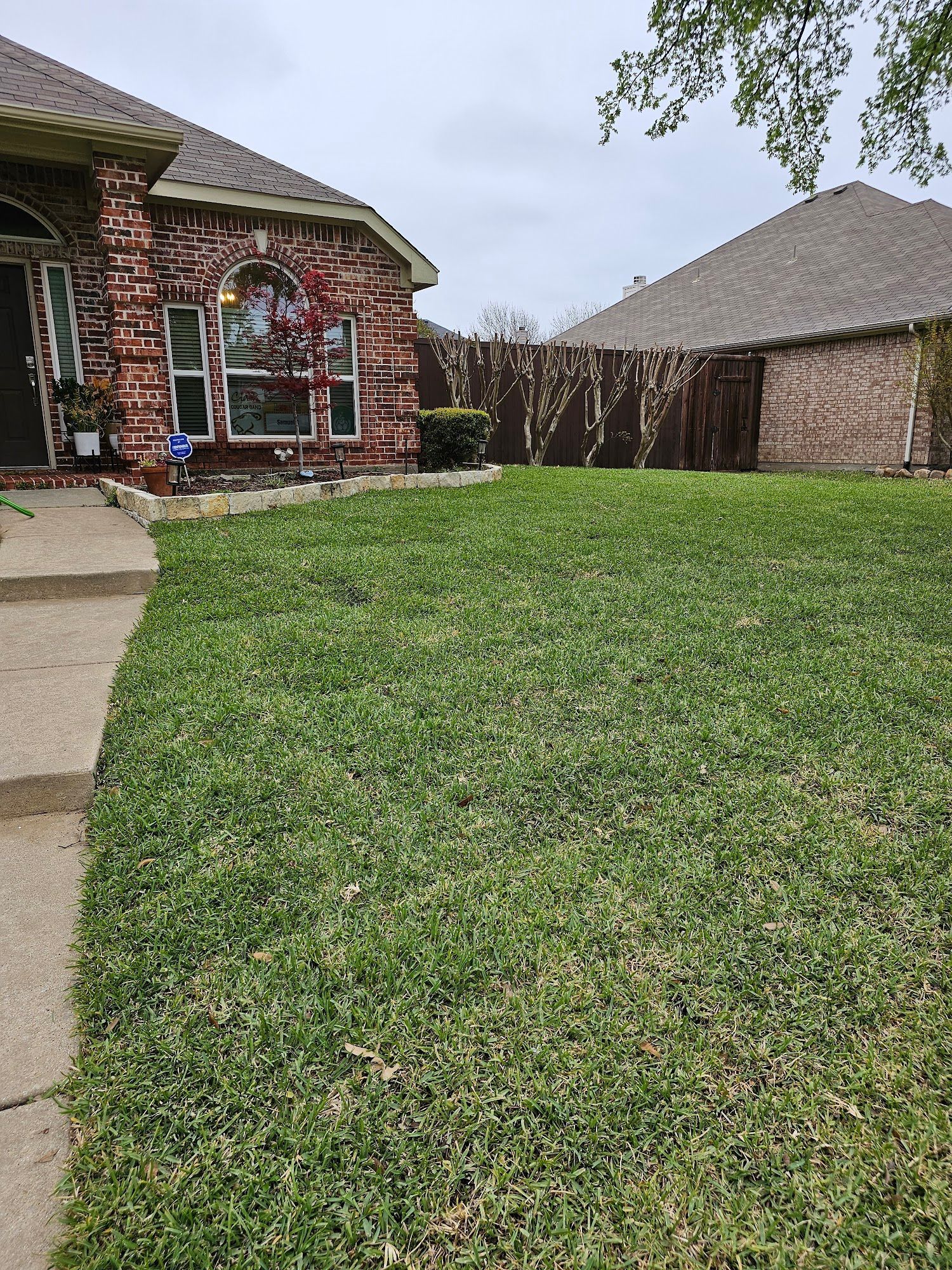 Green lawn in front of a brick house with brown roof and a wooden fence on a cloudy day.