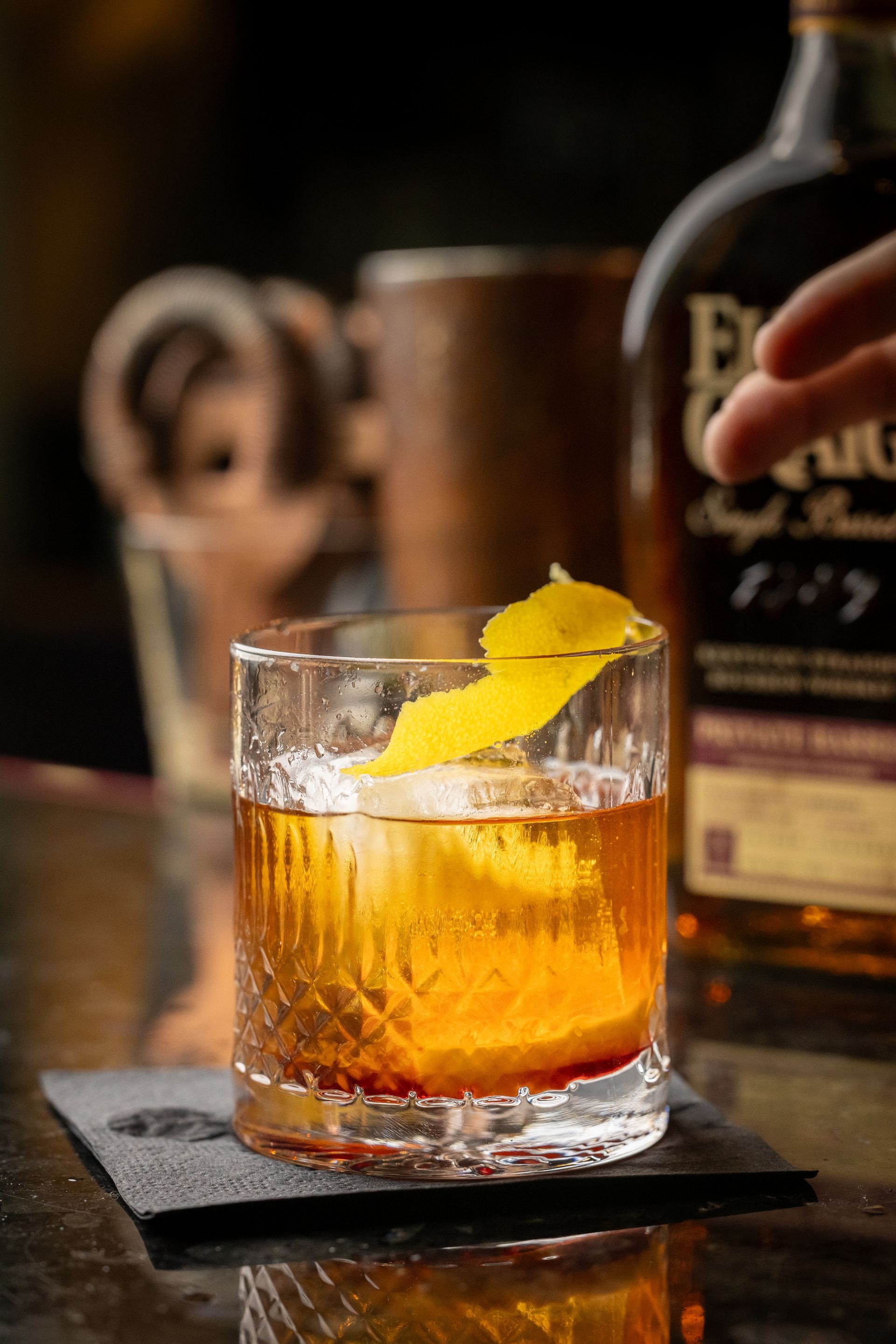 A person is holding a glass of whiskey on a coaster next to a bottle of whiskey.