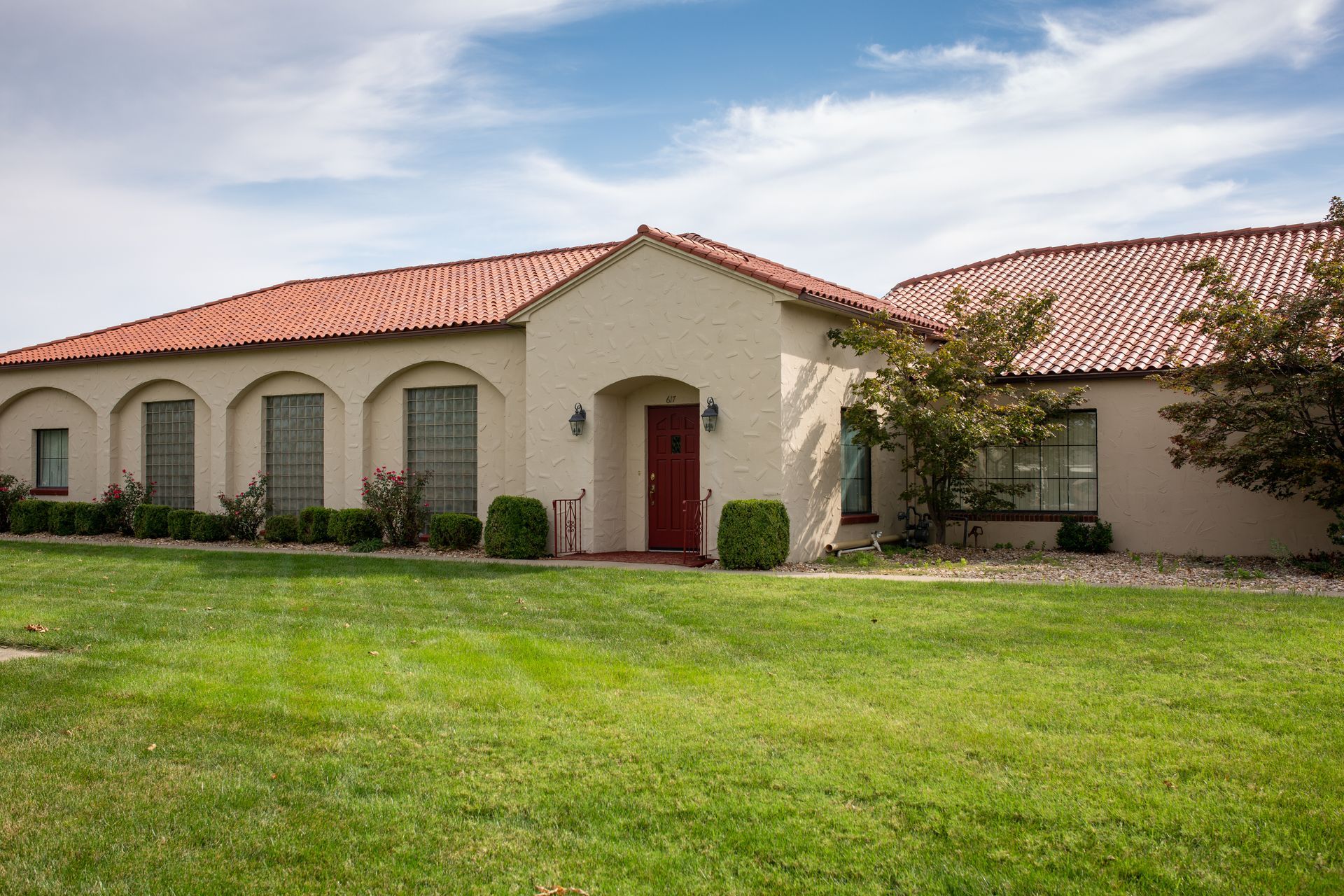 Single-story beige house with red tile roof, arched windows, and a green lawn in front